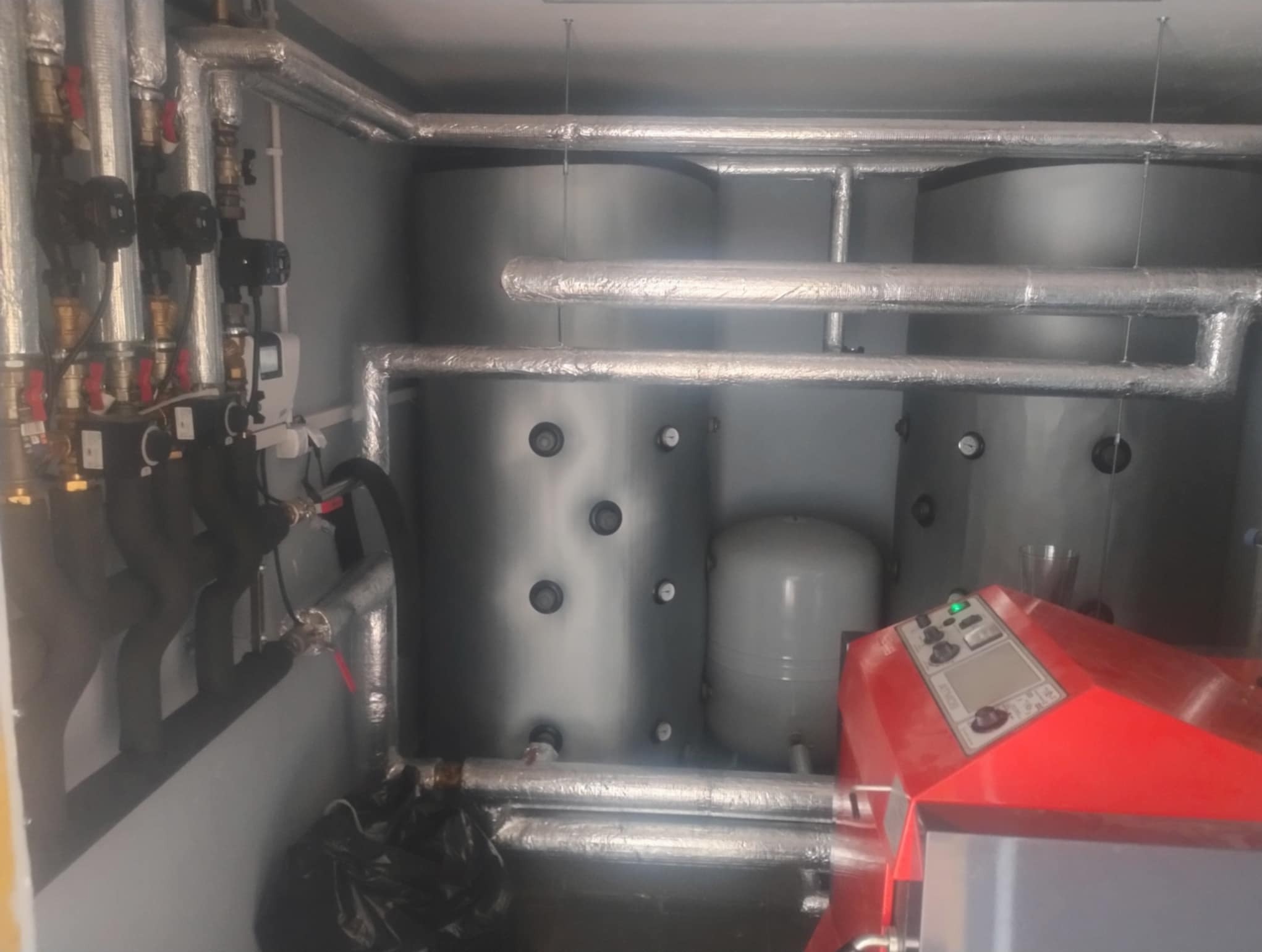 Złożona instalacja grzewcza z czerwonym piecem, zbiornikami akumulacyjnymi i izolowanymi rurami w pomieszczeniu technicznym. Widoczne liczne zawory i czujniki.