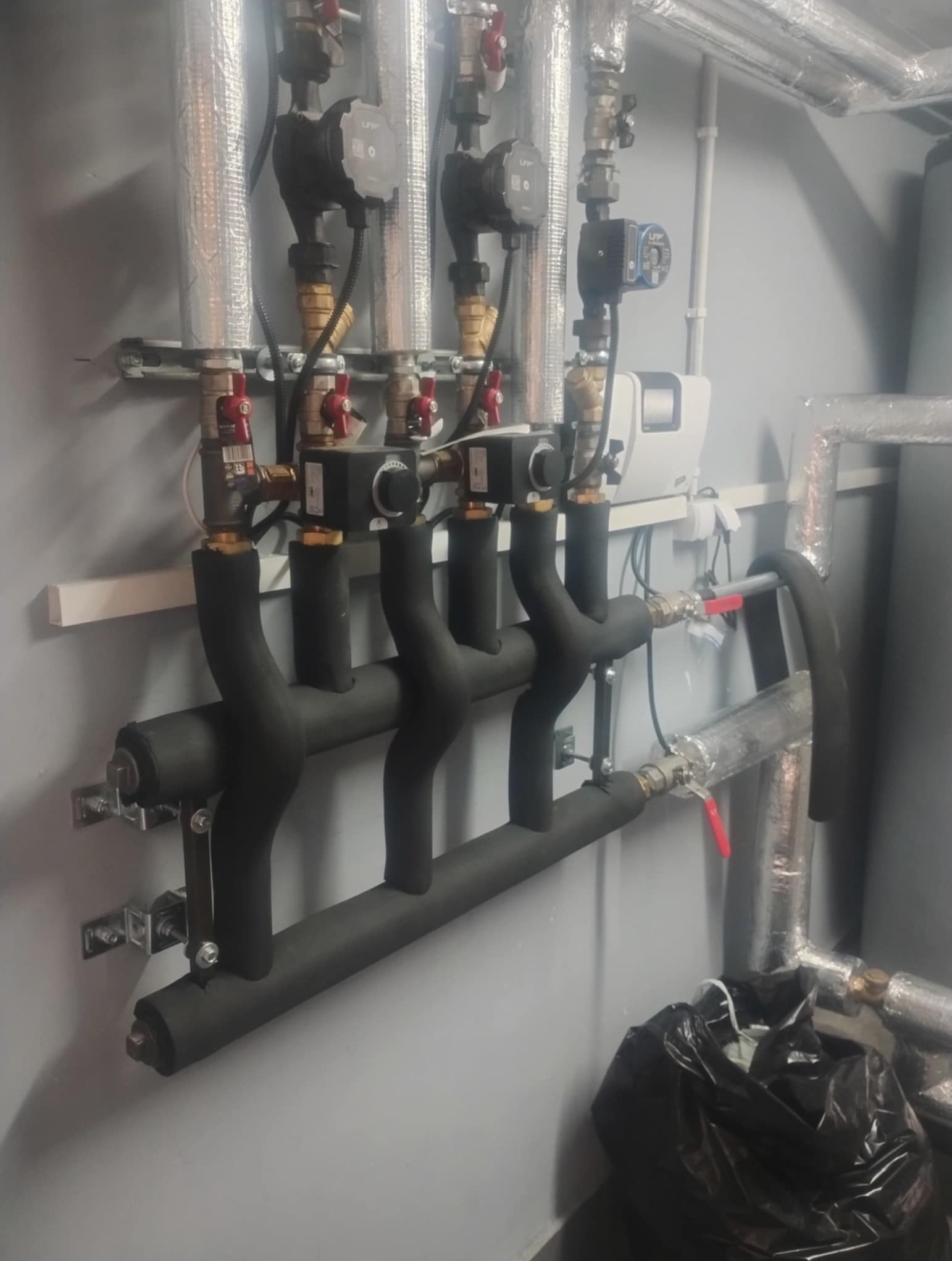 Rozdzielacz ogrzewania podłogowego z pompami obiegowymi, rurami w izolacji termicznej i zaworami z czerwonymi pokrętłami, zamontowany na szarej ścianie.