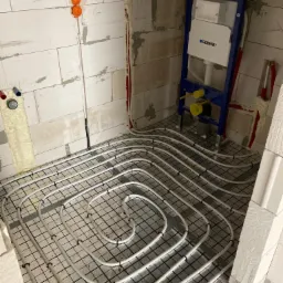 Białe rury ogrzewania podłogowego ułożone spiralnie na czarnej siatce w surowym wnętrzu łazienki, widoczny stelaż podtynkowy Geberit i instalacje w ścianach z betonu komórkowego.
