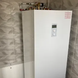 Biały piec elektryczny Kospel z podłączonymi rurami miedzianymi i plastikowymi w łazience wyłożonej szarymi płytkami.