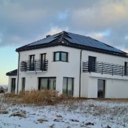 Nowoczesny, dwupiętrowy dom z białymi ścianami i panelami słonecznymi na dachu, widziany w zimowym krajobrazie z resztkami śniegu i suchą trawą na pierwszym planie.