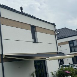SMARTEKS Sp. z o.o. - Nowoczesny dom z rozłożoną markizą w kolorze grafitowym, widok z dołu. Na ścianie widoczny monitoring. W tle drugi budynek i samochody.