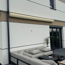 SMARTEKS Sp. z o.o. - Zamknięta, beżowa markiza zwinięta na ścianie nowoczesnego domu z szarą sofą ogrodową i czarnym stolikiem na tarasie z desek kompozytowych.