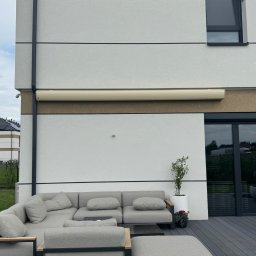 SMARTEKS Sp. z o.o. - Elegancka markiza zwinięta na nowoczesnym domu z szarą sofą na tarasie. Widok z zewnątrz, jasne ściany, czarne ramy okienne i drzwi.