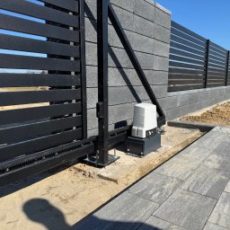SMARTEKS Sp. z o.o. - Nowoczesna, przesuwna brama z czarnymi, poziomymi panelami i szarym, betonowym ogrodzeniem. Widoczny napęd bramy i fragment podjazdu z szarej kostki brukowej.