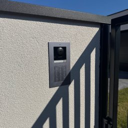 SMARTEKS Sp. z o.o. - Szary wideodomofon Hikvision zamontowany na elewacji budynku, widoczne elementy bramy i cień. Błękitne niebo w tle. Detal instalacji.