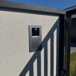 Szary wideodomofon Hikvision zamontowany na elewacji budynku, widoczne elementy bramy i cień. Błękitne niebo w tle. Detal instalacji.