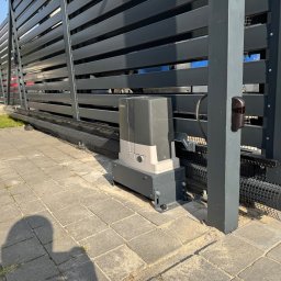SMARTEKS Sp. z o.o. - Szara, przesuwna brama z automatyką, widoczny napęd i fotokomórka. Brama z poziomymi panelami, zamontowana na posesji z kostki brukowej.