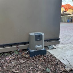 SMARTEKS Sp. z o.o. - Szary automat do bramy Somfy, zamontowany przy brązowej bramie przesuwnej, na tle kostki brukowej i budynku z czerwoną dachówką. Widoczne liście i ziemia.