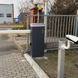 Szary automat do bram Came z czerwoną listwą LED, kamera BCS na słupku i fragment bramy na tle budynków i drzew.