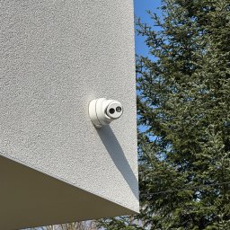 SMARTEKS Sp. z o.o. - Biała kamera monitoringu, zamontowana na elewacji budynku o jasnej strukturze, z iglastym drzewem w tle i fragmentem nieba.