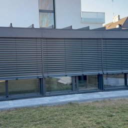 SMARTEKS Sp. z o.o. - Żaluzje fasadowe w kolorze antracytowym zamontowane na parterze nowoczesnego budynku. Widoczne trzy okna z opuszczonymi żaluzjami, fragment trawnika i elewacji.
