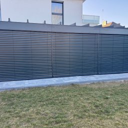 SMARTEKS Sp. z o.o. - Zewnętrzne żaluzje fasadowe w kolorze grafitowym, zamontowane w nowoczesnej pergoli przy domu z kostki brukowej i trawnikiem. Widok z poziomu gruntu, słoneczny dzień.