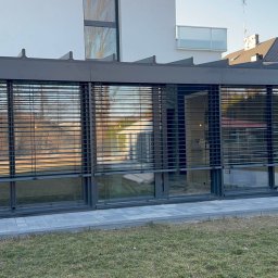 SMARTEKS Sp. z o.o. - Żaluzje fasadowe w nowoczesnej zabudowie, z odbiciem osoby w jednej z szyb. Widok z zewnątrz na tle zieleni i kostki brukowej.