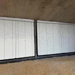 Dwie białe rolety zewnętrzne zamontowane w surowym stanie budynku, widoczne cegły i betonowy strop. Rolety w pozycji zamkniętej, ochrona przed słońcem i włamaniem.