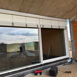 SMARTEKS Sp. z o.o. - Zamknięte białe żaluzje fasadowe zamontowane w oknie budynku w trakcie budowy. Widoczne cegły, narzędzia, skrzynki narzędziowe. Ujęcie z dołu, z zewnątrz.