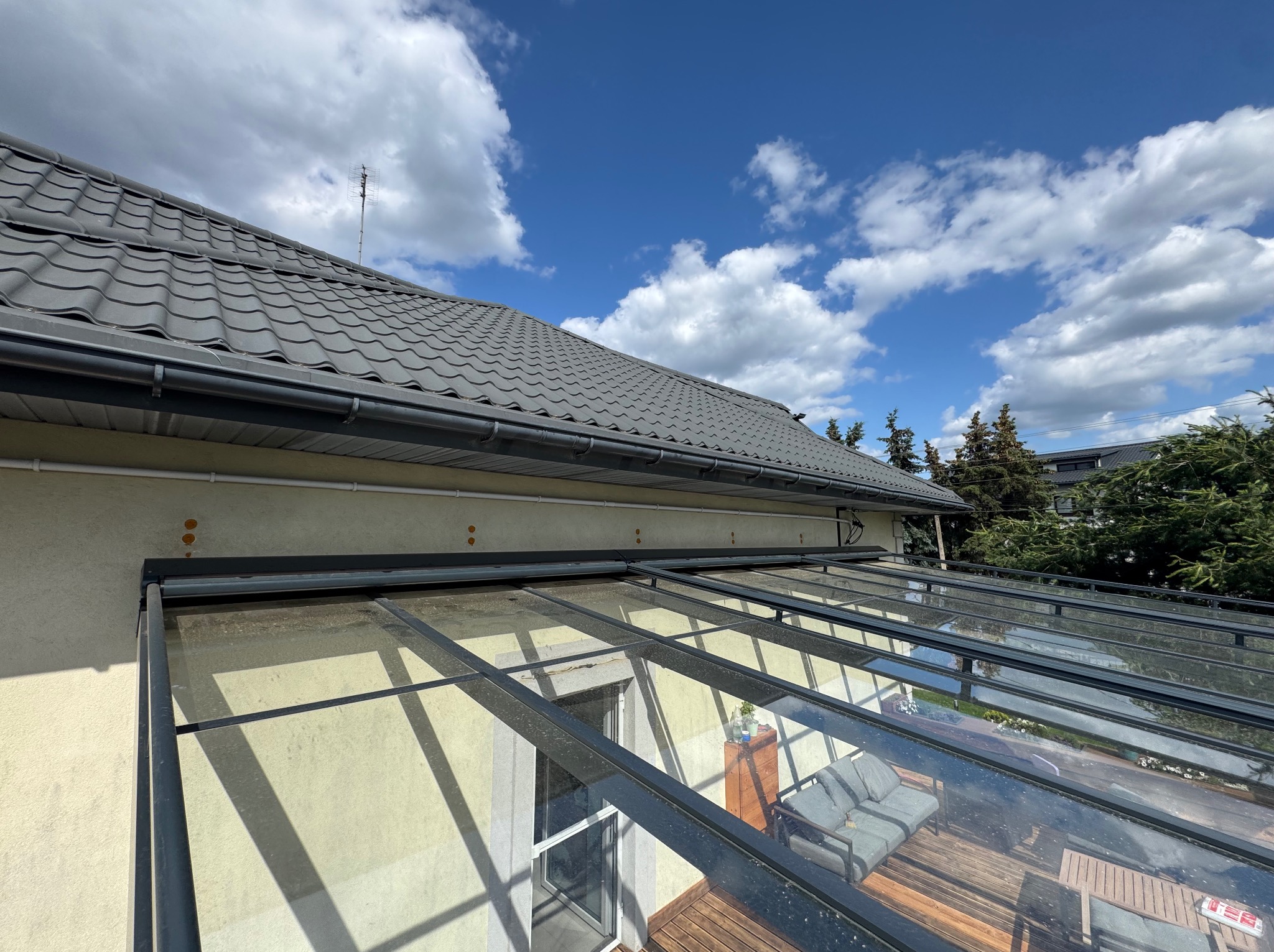 Szklana pergola metalowa przylegająca do budynku, widok z dołu na konstrukcję i dach, z meblami ogrodowymi widocznymi wewnątrz, błękitne niebo w tle.