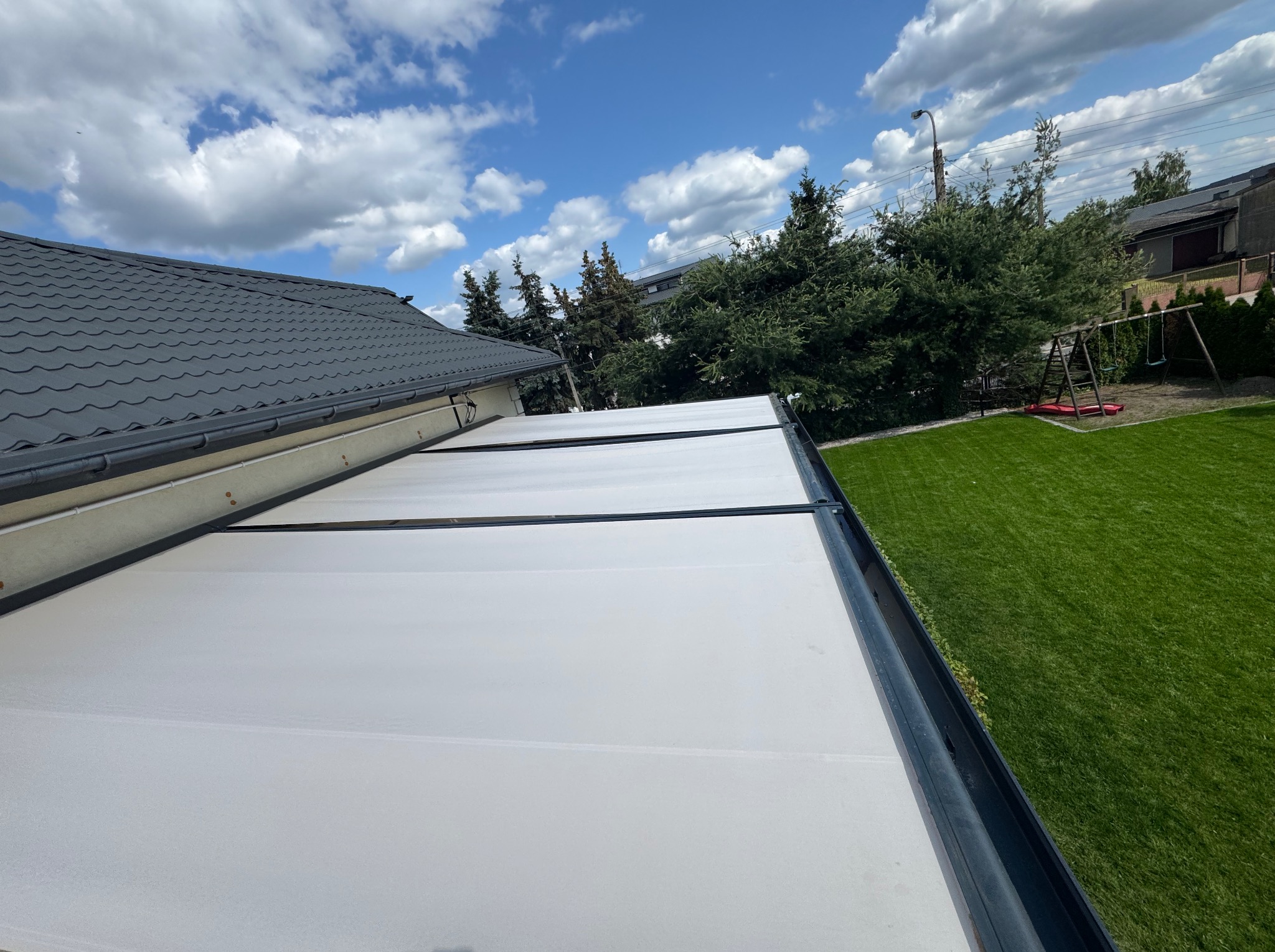 Nowoczesna pergola tarasowa z jasnym zadaszeniem, widok z dołu na tle błękitnego nieba i zielonego trawnika. Widoczne elementy konstrukcyjne i zarys dachu budynku.