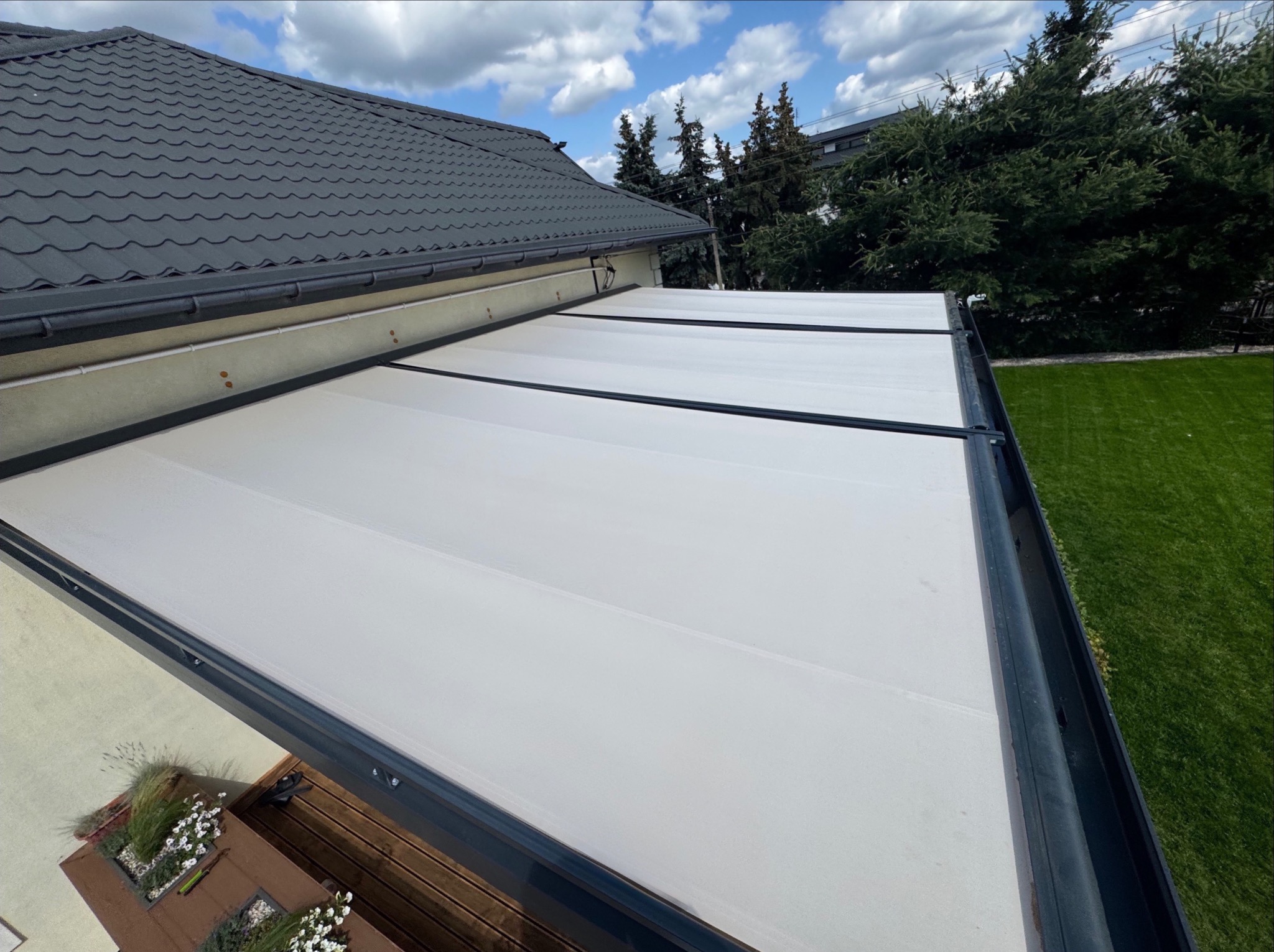 Zewnętrzna, rozsuwana pergola z białym, materiałowym dachem, zamontowana przy domu z szarą dachówką. Widok z góry, w tle zieleń drzew i trawnika.