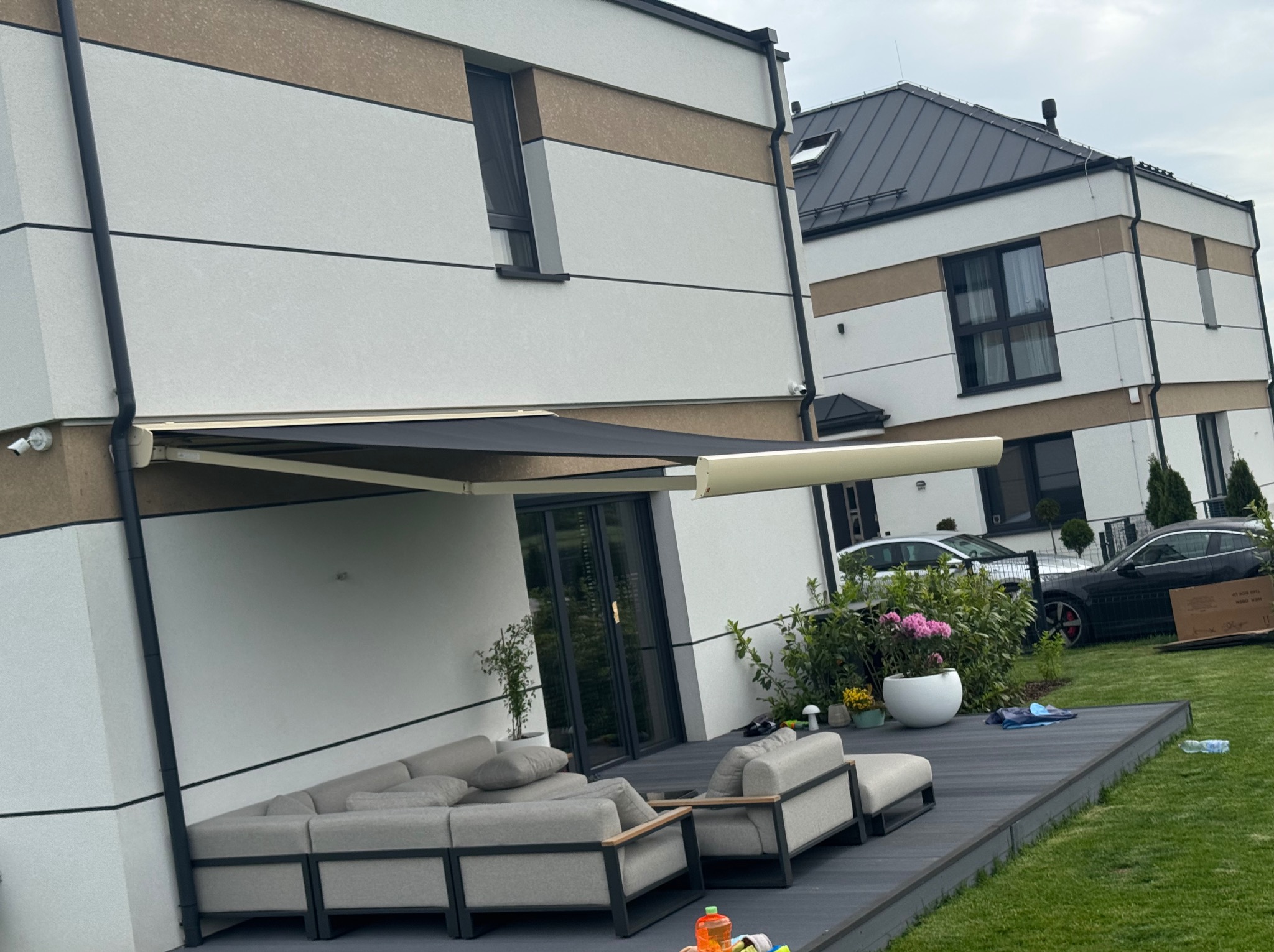 Elegancka markiza tarasowa w kolorze antracytowym, zamontowana przy nowoczesnym domu z szarym narożnikiem na drewnianym tarasie. Widok na zadbany ogród i zaparkowane samochody.
