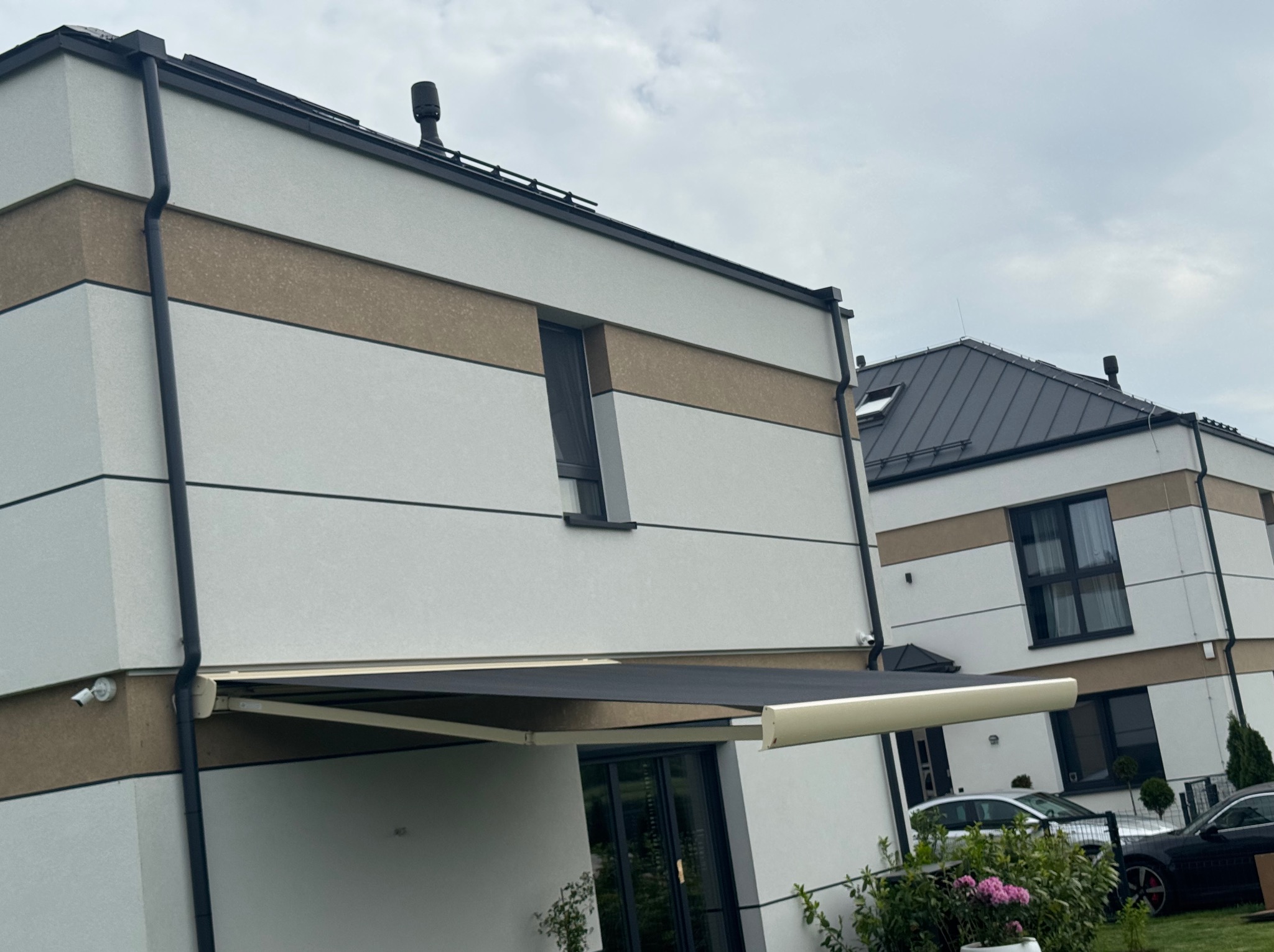 Nowoczesny dom z rozłożoną markizą w kolorze grafitowym, widok z dołu. Na ścianie widoczny monitoring. W tle drugi budynek i samochody.