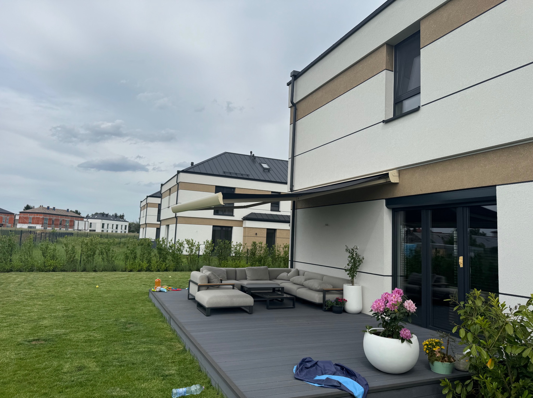 Nowoczesny taras z szarą sofą i pergolą metalową przy domu w jasnych kolorach. Na trawniku leżą zabawki.