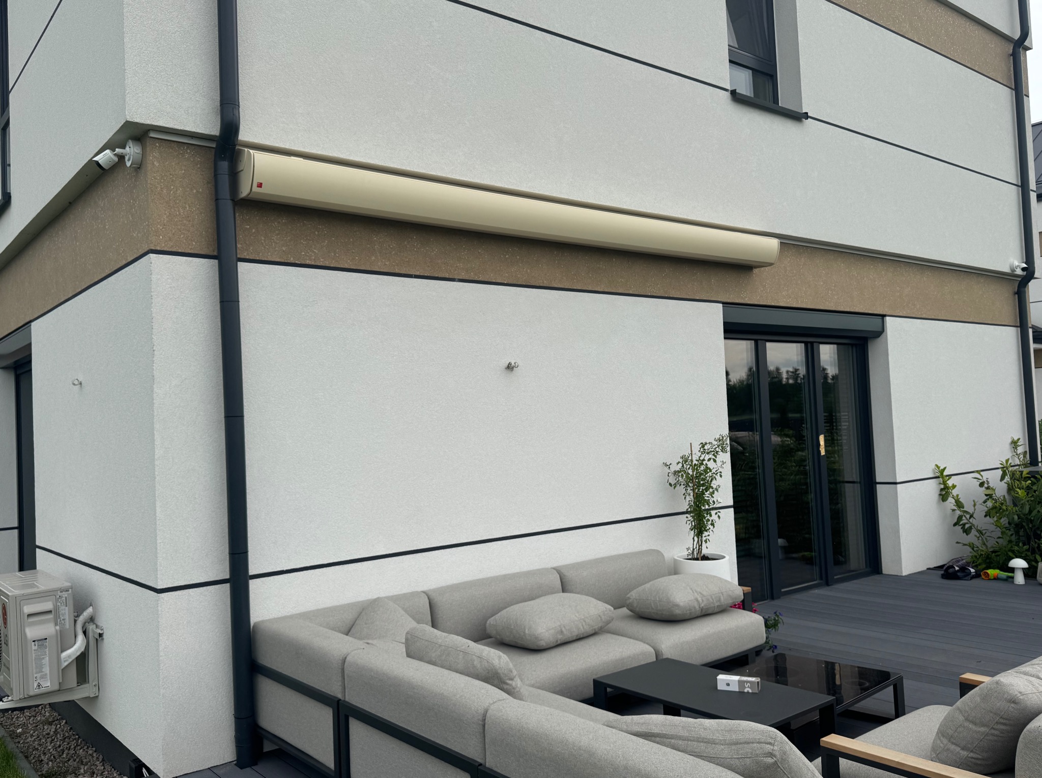 Zamknięta, beżowa markiza zwinięta na ścianie nowoczesnego domu z szarą sofą ogrodową i czarnym stolikiem na tarasie z desek kompozytowych.
