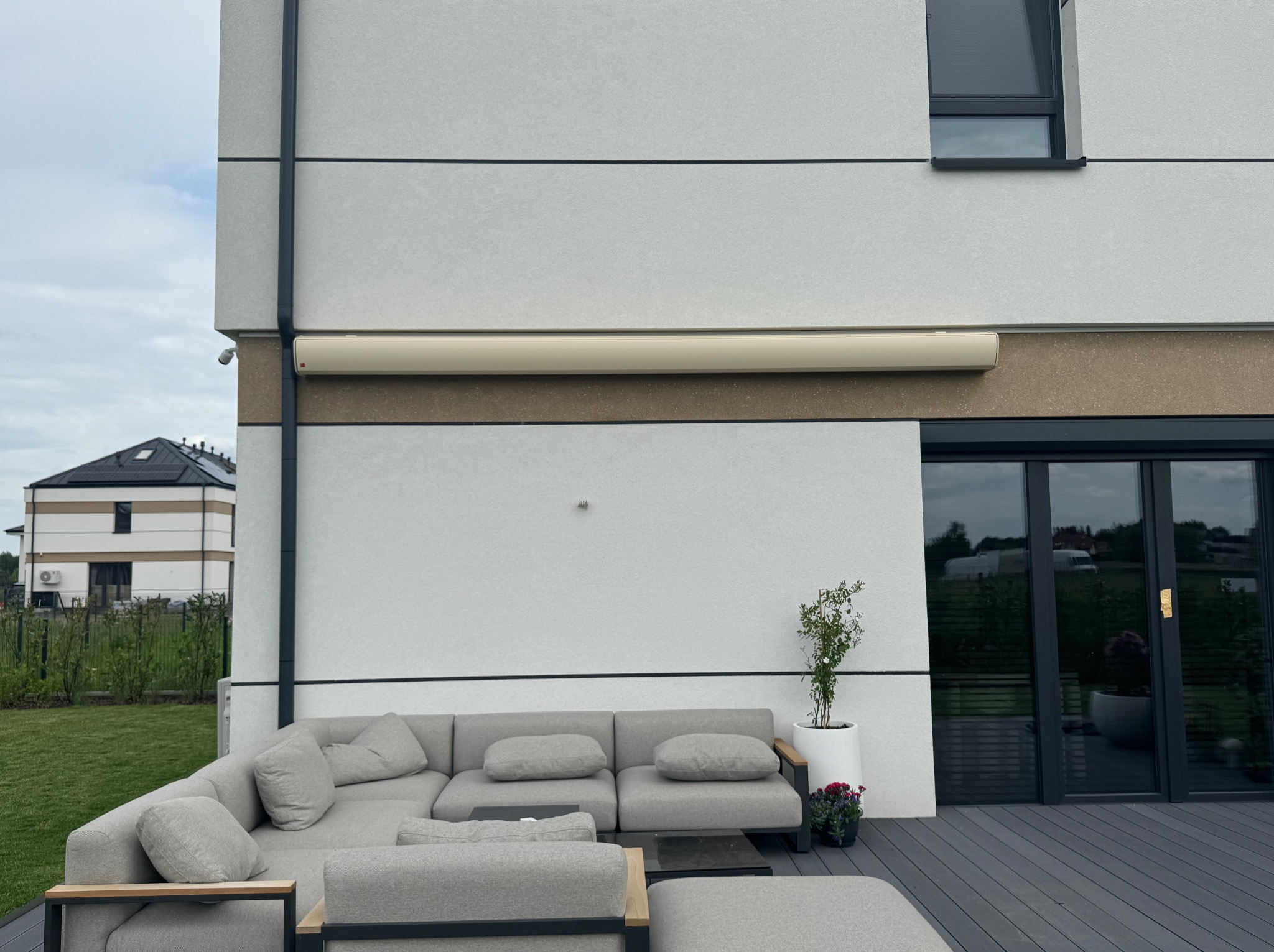 Elegancka markiza zwinięta na nowoczesnym domu z szarą sofą na tarasie. Widok z zewnątrz, jasne ściany, czarne ramy okienne i drzwi.