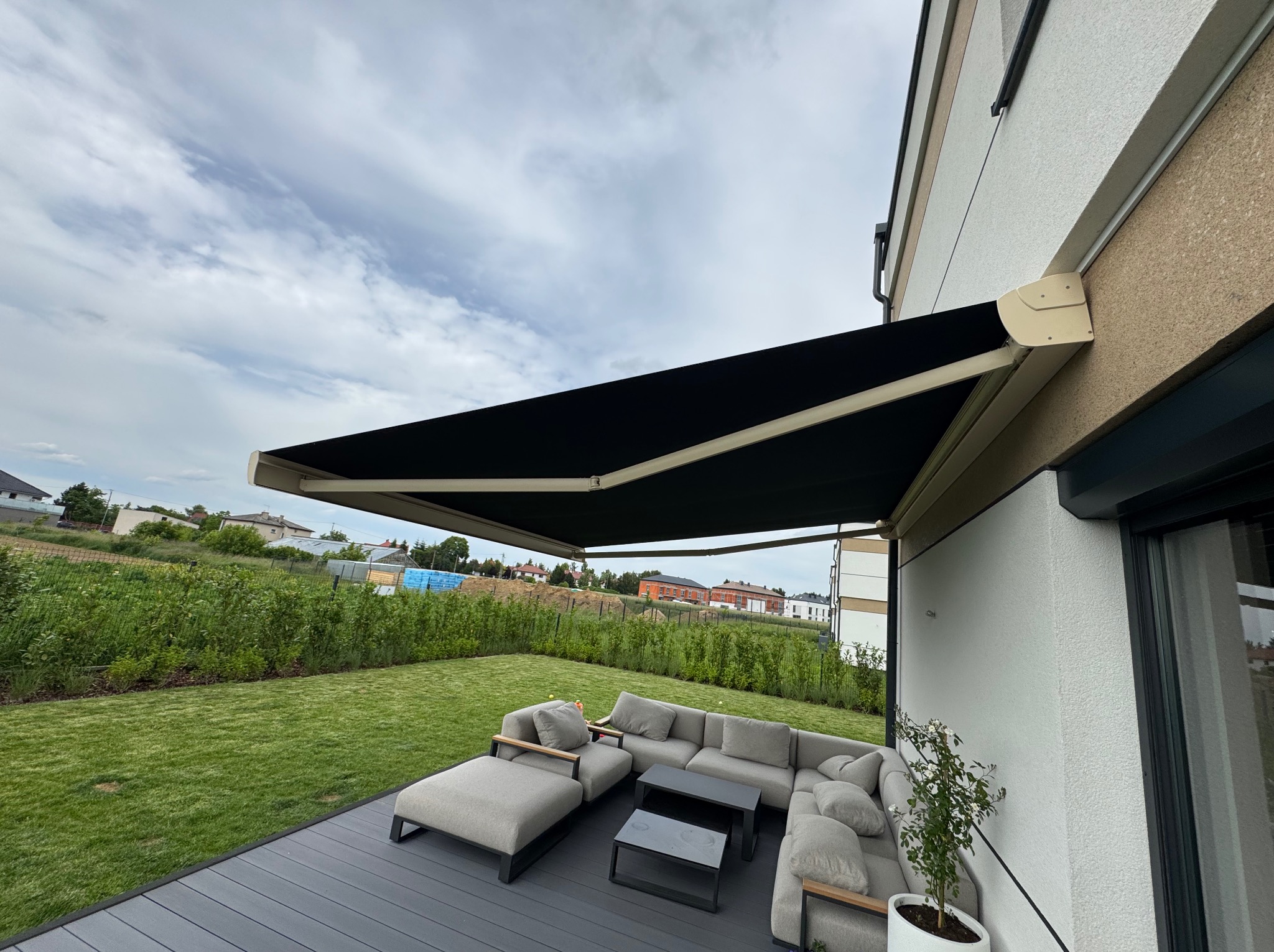 Czarna markiza tarasowa zamontowana przy nowoczesnym domu z jasną elewacją. Na tarasie szare meble ogrodowe i zadbany trawnik w tle.