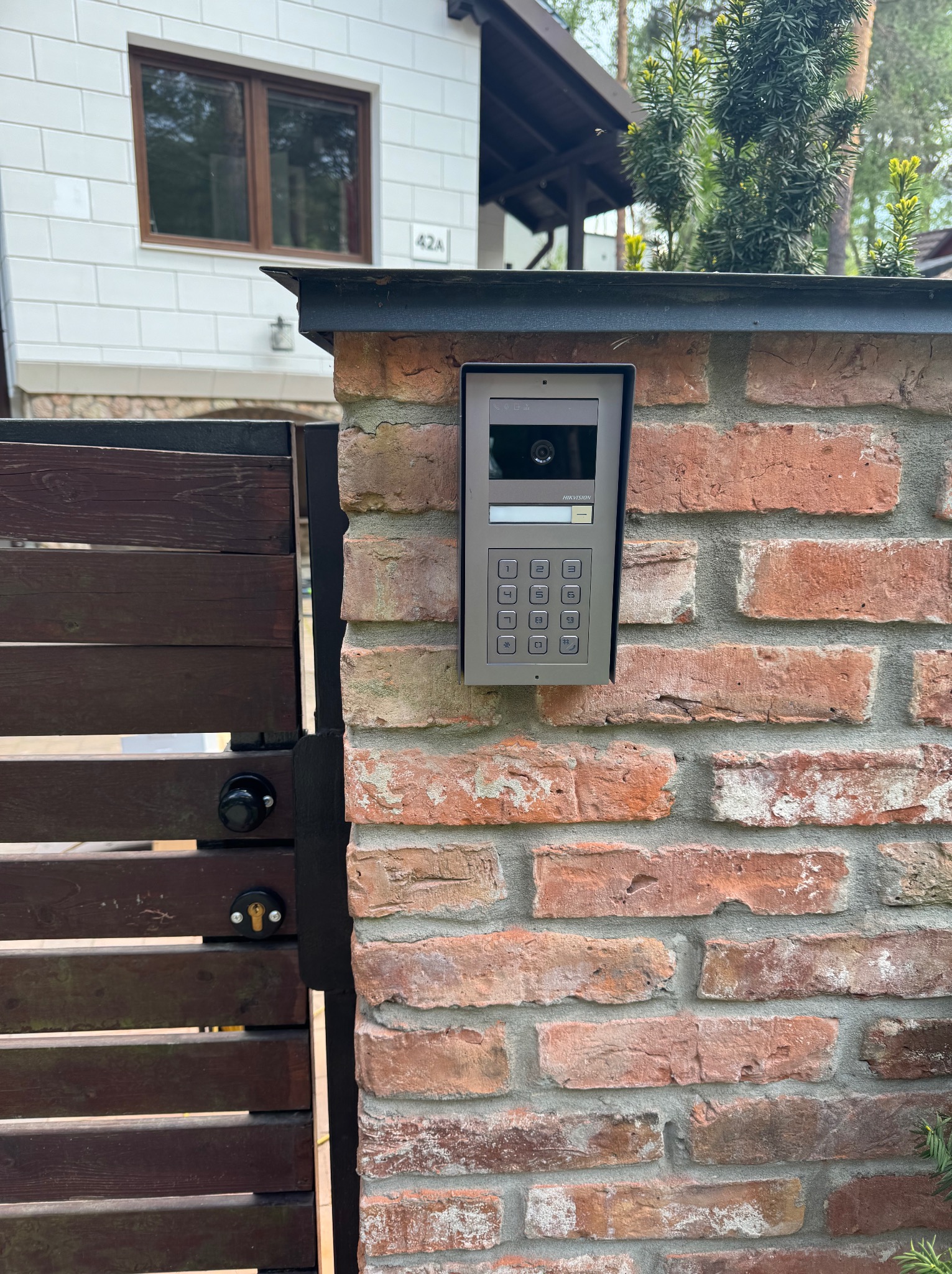 Szary wideofon Hikvision zamontowany na ceglanym słupku ogrodzenia, z widoczną bramą z drewnianych paneli i fragmentem elewacji budynku.