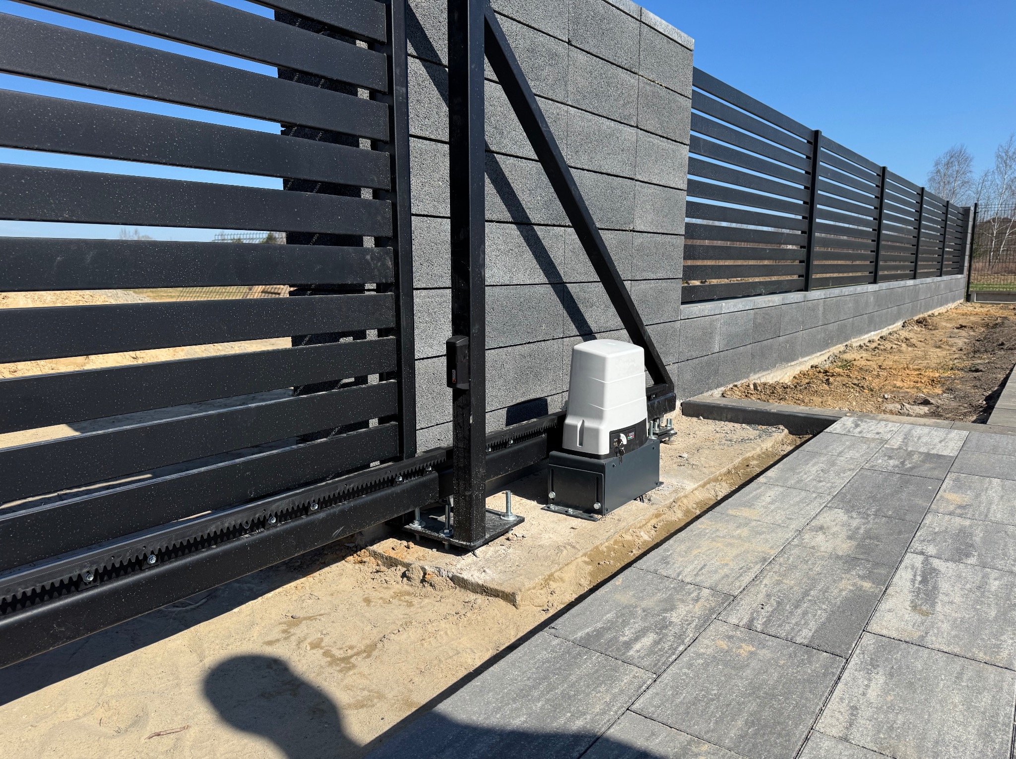 Nowoczesna, przesuwna brama z czarnymi, poziomymi panelami i szarym, betonowym ogrodzeniem. Widoczny napęd bramy i fragment podjazdu z szarej kostki brukowej.