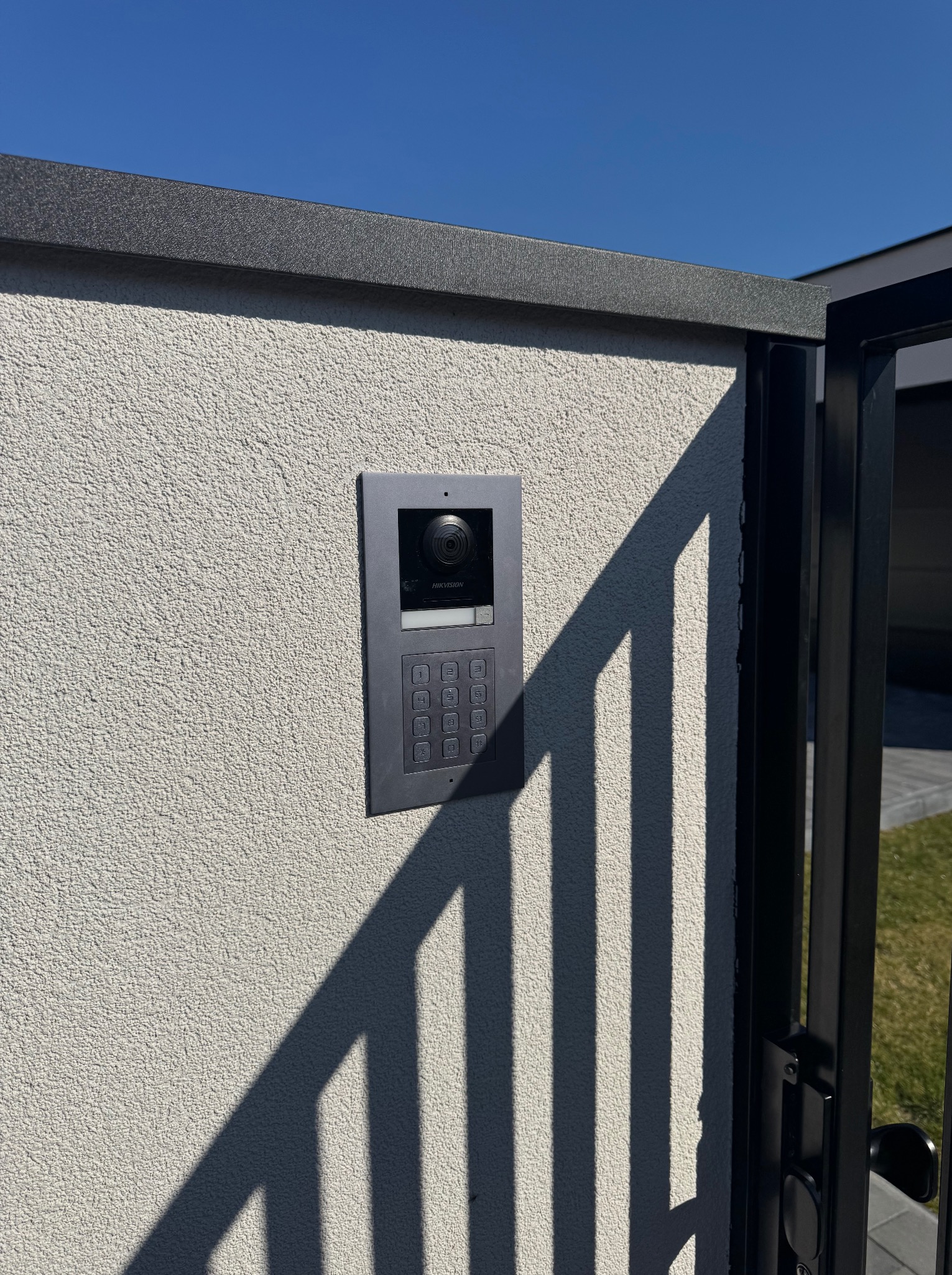 Szary wideodomofon Hikvision zamontowany na elewacji budynku, widoczne elementy bramy i cień. Błękitne niebo w tle. Detal instalacji.