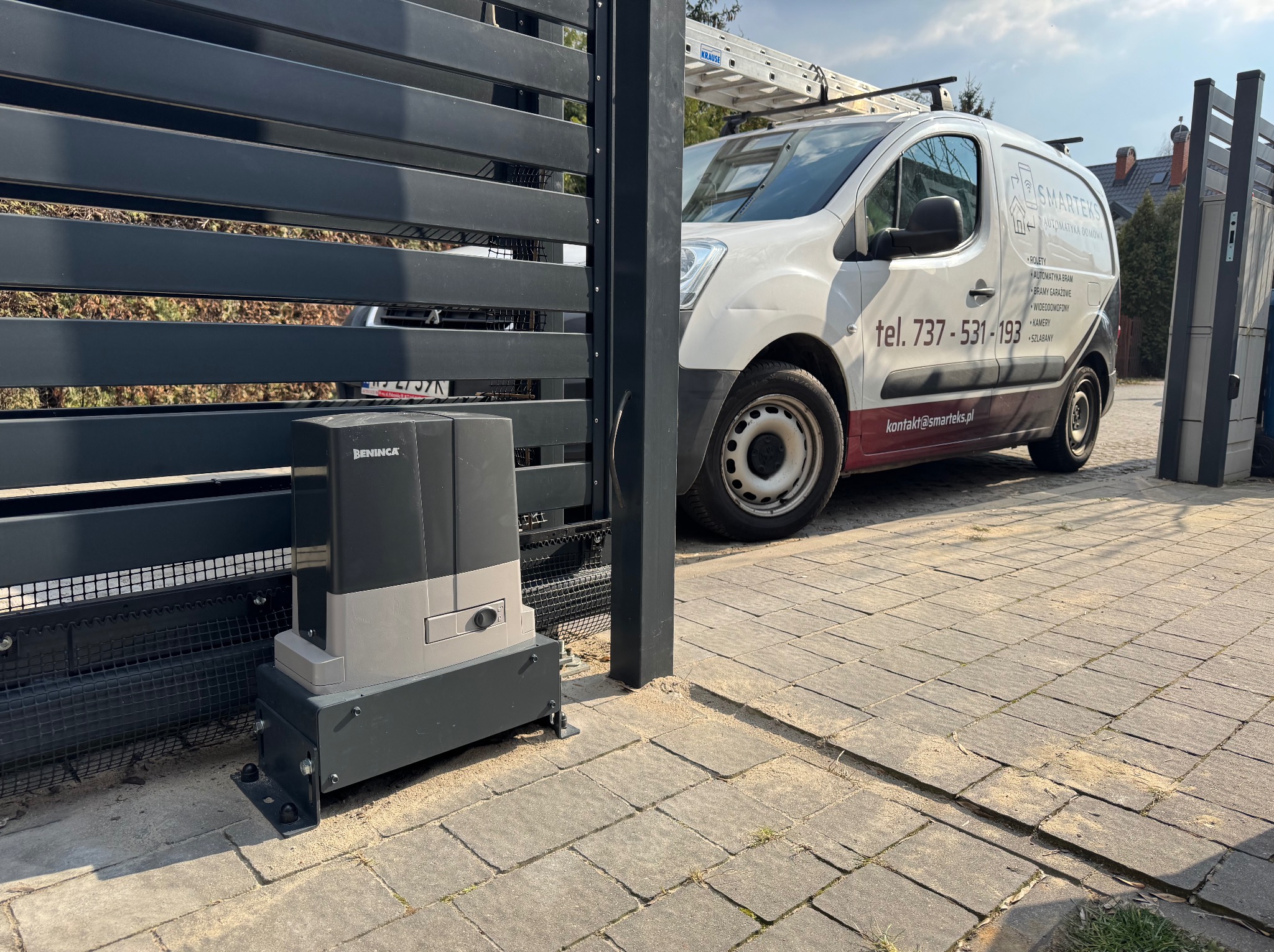 Zamontowany napęd do bramy przesuwnej Beninca przy nowej, poziomej bramie. W tle biały samochód dostawczy z logo firmy i drabiną na dachu.