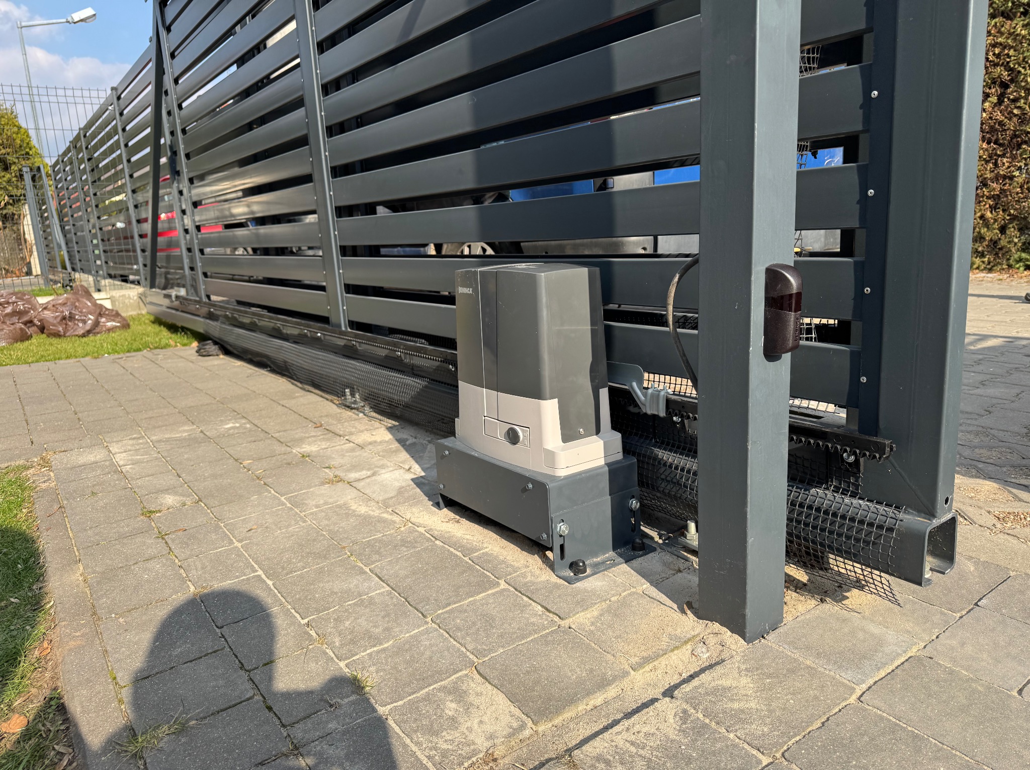 Szara, przesuwna brama z automatyką, widoczny napęd i fotokomórka. Brama z poziomymi panelami, zamontowana na posesji z kostki brukowej.