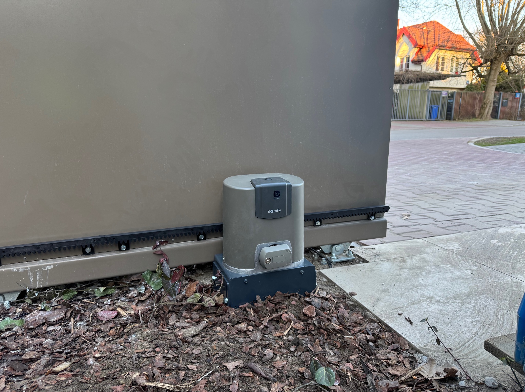 Szary automat do bramy Somfy, zamontowany przy brązowej bramie przesuwnej, na tle kostki brukowej i budynku z czerwoną dachówką. Widoczne liście i ziemia.