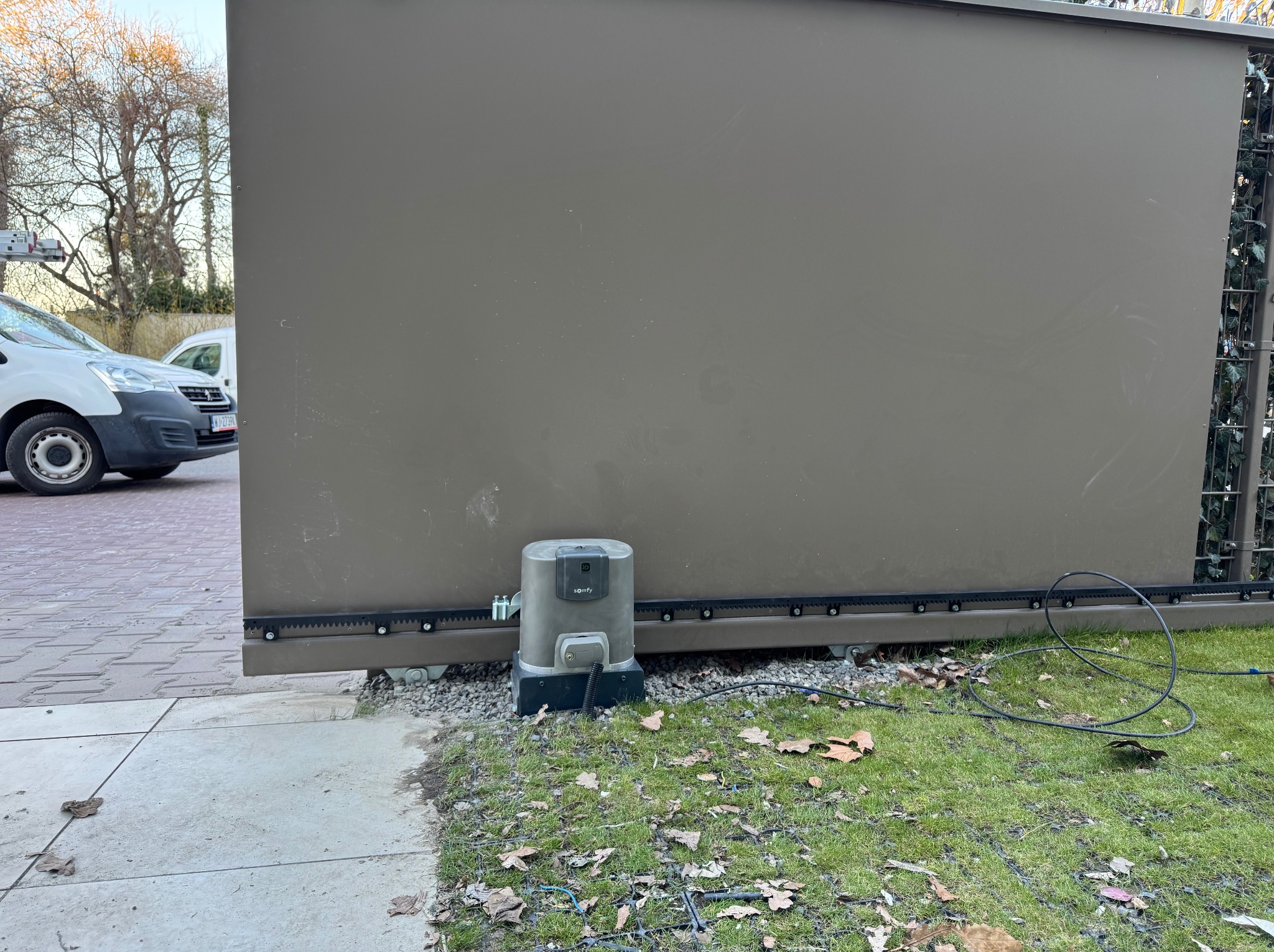 Szara, przesuwna brama z automatyką Somfy, widoczna na tle trawnika i chodnika. Fragment białego samochodu w tle. Brama częściowo zasłania metalową konstrukcję z roślinami.