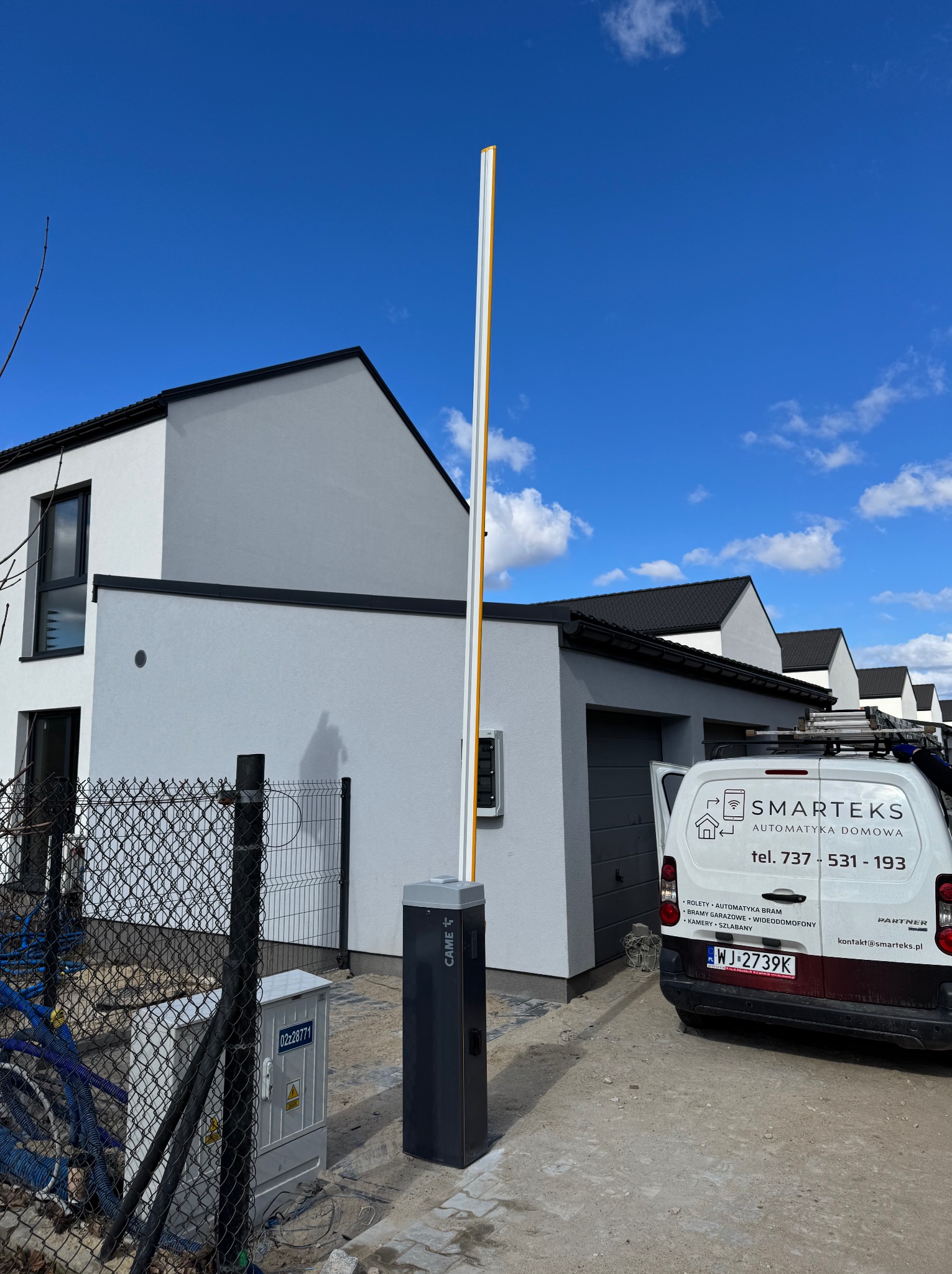Nowoczesny szlaban automatyczny CAME przed domem, z widocznym samochodem firmowym Smarteks z listą usług: rolety, bramy, kamery. Błękitne niebo w tle.