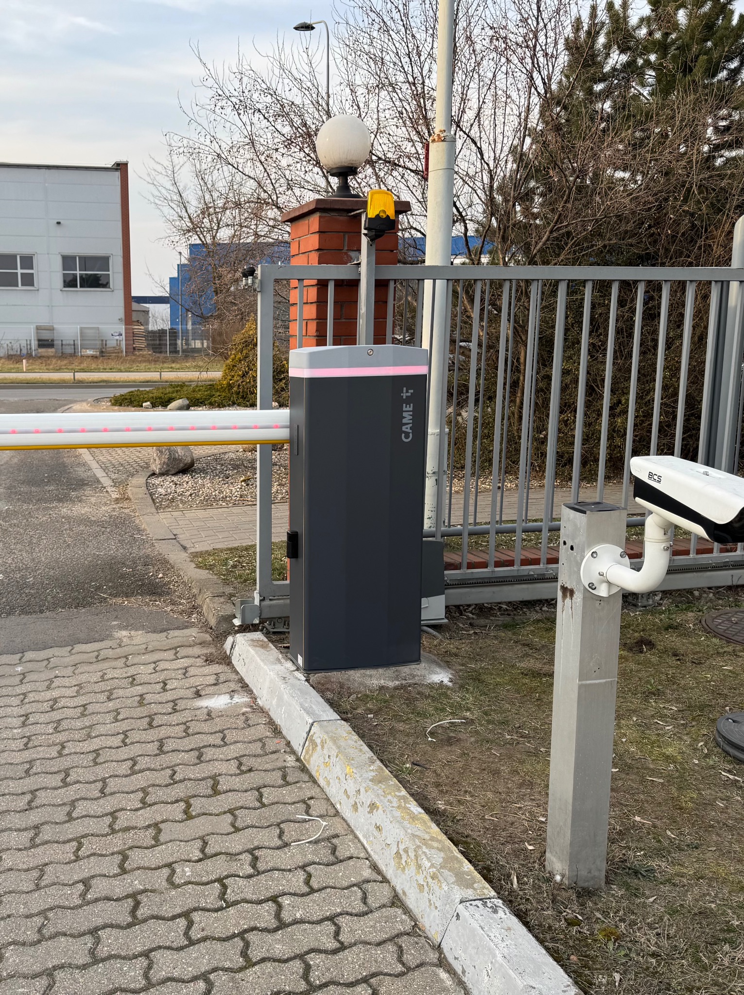 Szary automat do bram Came z czerwoną listwą LED, kamera BCS na słupku i fragment bramy na tle budynków i drzew.