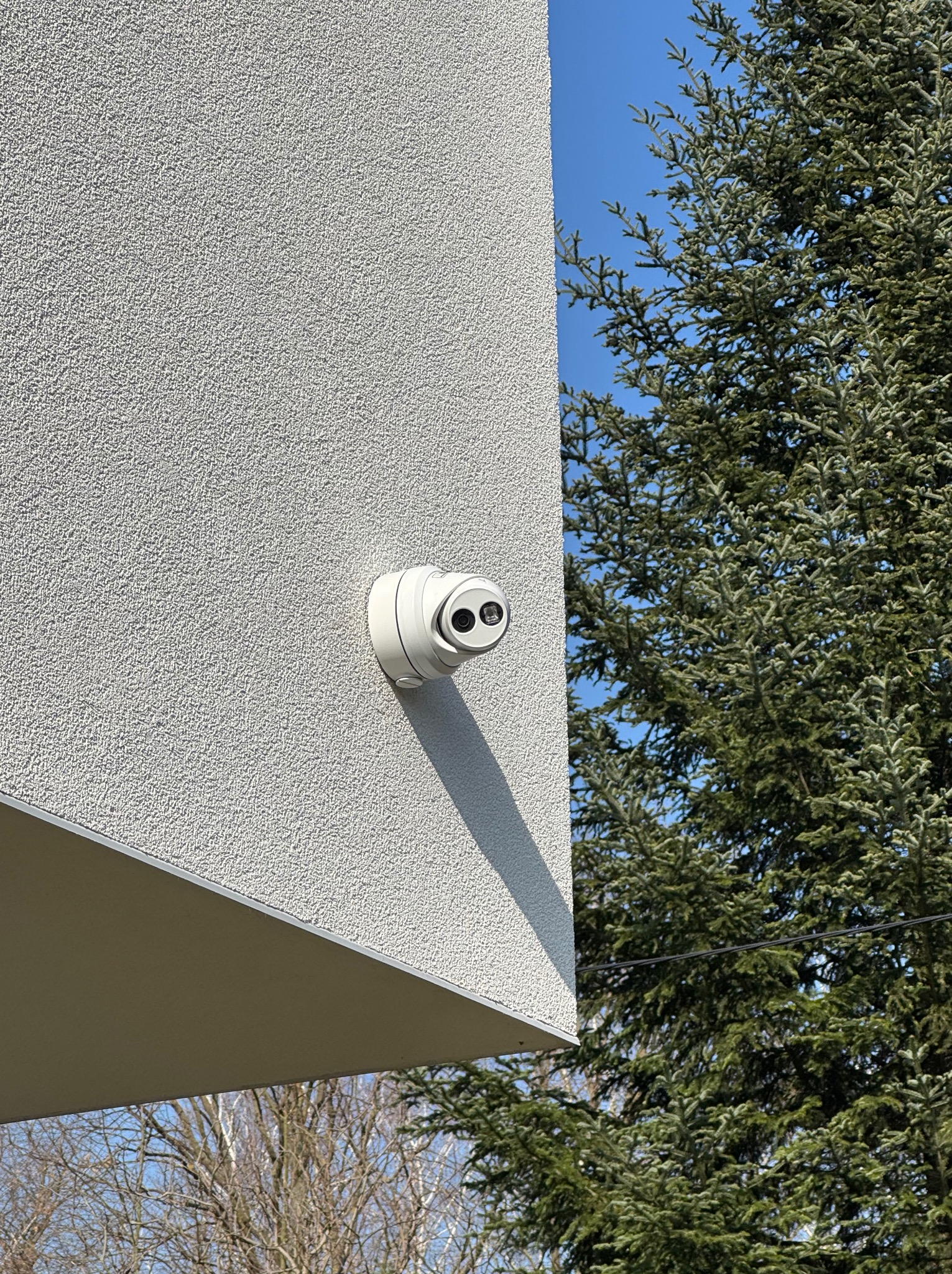 Biała kamera monitoringu, zamontowana na elewacji budynku o jasnej strukturze, z iglastym drzewem w tle i fragmentem nieba.