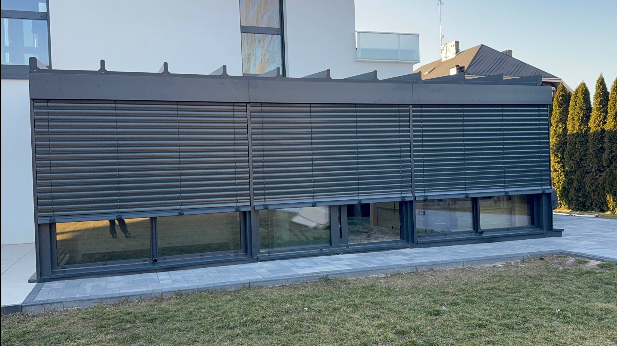Żaluzje fasadowe w kolorze antracytowym zamontowane na parterze nowoczesnego budynku. Widoczne trzy okna z opuszczonymi żaluzjami, fragment trawnika i elewacji.