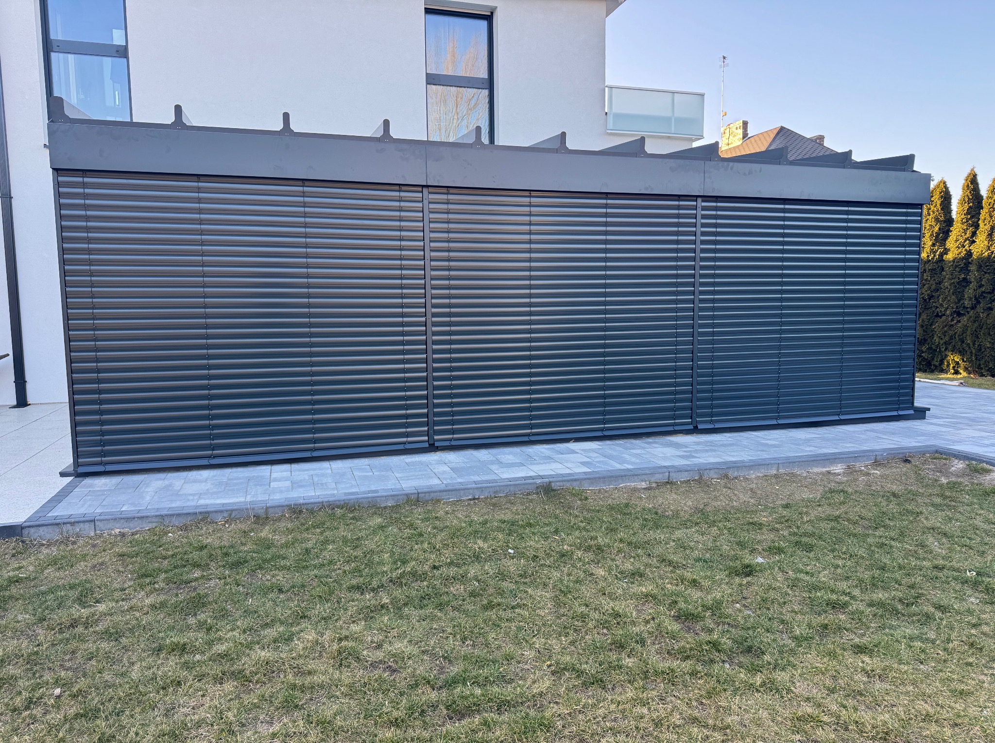 Zewnętrzne żaluzje fasadowe w kolorze grafitowym, zamontowane w nowoczesnej pergoli przy domu z kostki brukowej i trawnikiem. Widok z poziomu gruntu, słoneczny dzień.