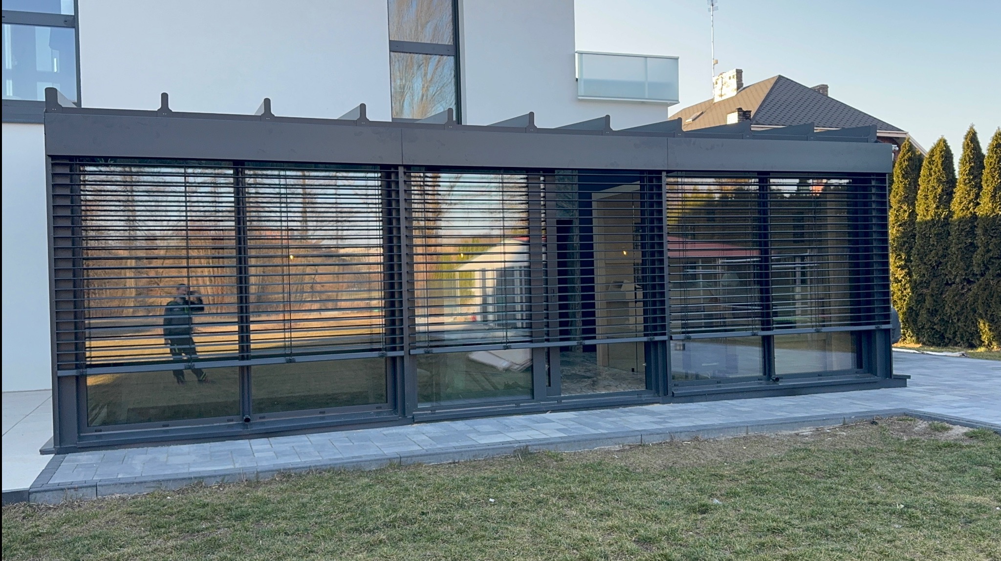 Żaluzje fasadowe w nowoczesnej zabudowie, z odbiciem osoby w jednej z szyb. Widok z zewnątrz na tle zieleni i kostki brukowej.