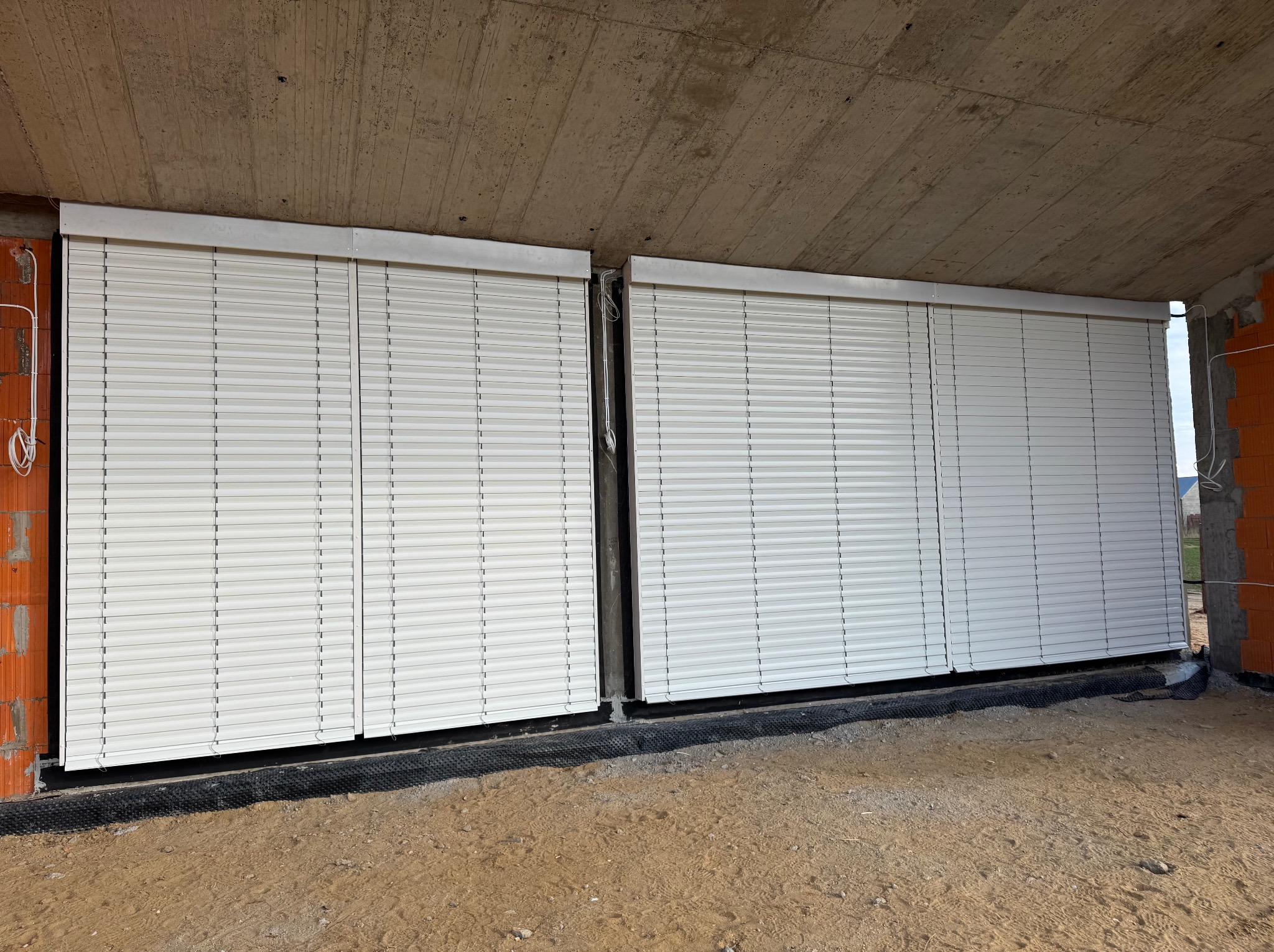 Dwie białe rolety zewnętrzne zamontowane w surowym stanie budynku, widoczne cegły i betonowy strop. Rolety w pozycji zamkniętej, ochrona przed słońcem i włamaniem.