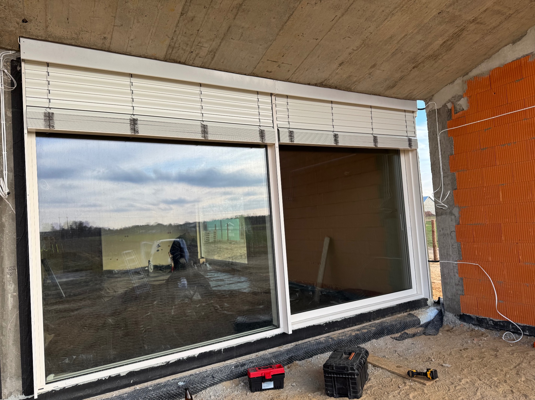 Zamknięte białe żaluzje fasadowe zamontowane w oknie budynku w trakcie budowy. Widoczne cegły, narzędzia, skrzynki narzędziowe. Ujęcie z dołu, z zewnątrz.