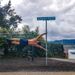 SPORTOWO SPÓŁKA Z OGRANICZONĄ ODPOWIEDZIALNOŚCIĄ - Mężczyzna w pozycji 'flaga' na słupie z tabliczką 'Street Workout' na tle jeziora i zieleni. Widoczne drabinki do ćwiczeń i łódź na wodzie.