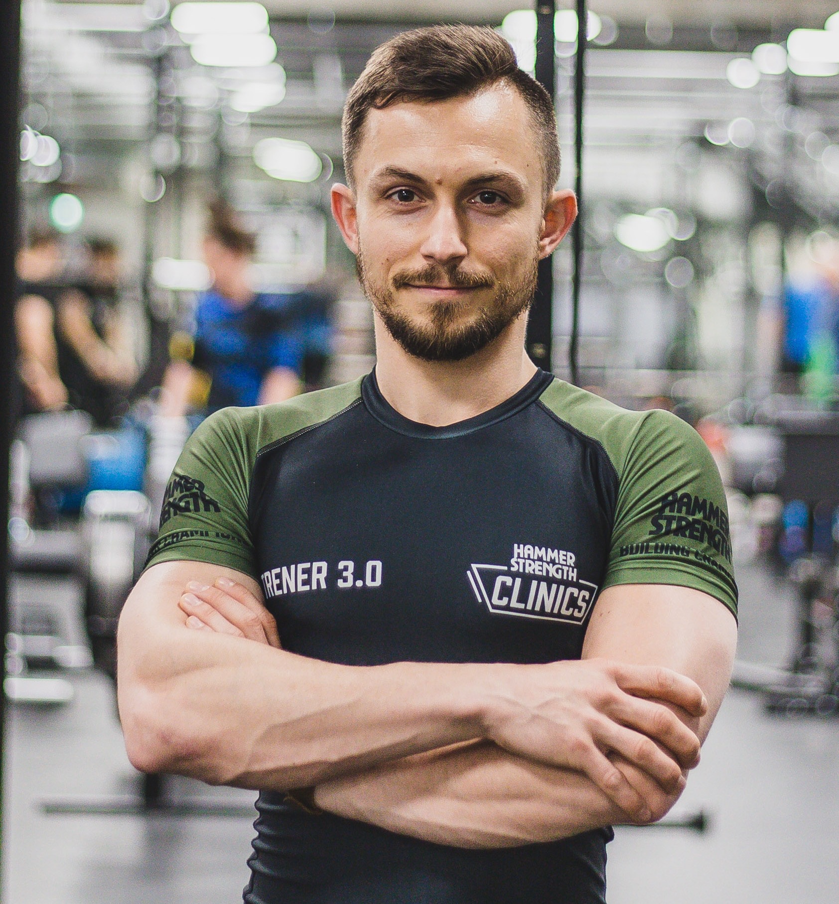Portret uśmiechniętego trenera personalnego w sportowej koszulce z logo Hammer Strength Clinics, na tle siłowni z ćwiczącymi osobami.