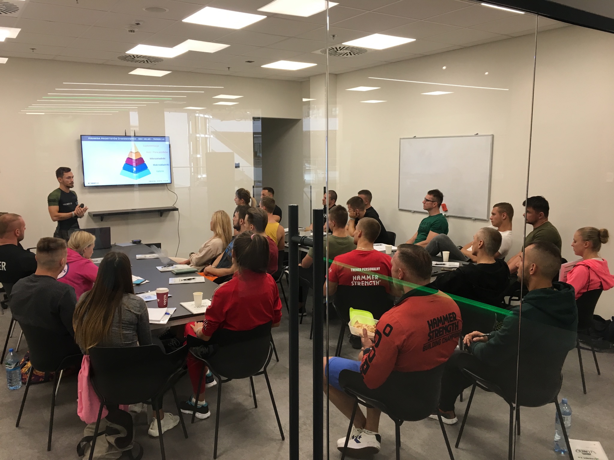 Prelegent omawia piramidę priorytetów żywieniowych na szkoleniu dla grupy osób, siedzących przy stole w nowoczesnej sali konferencyjnej.
