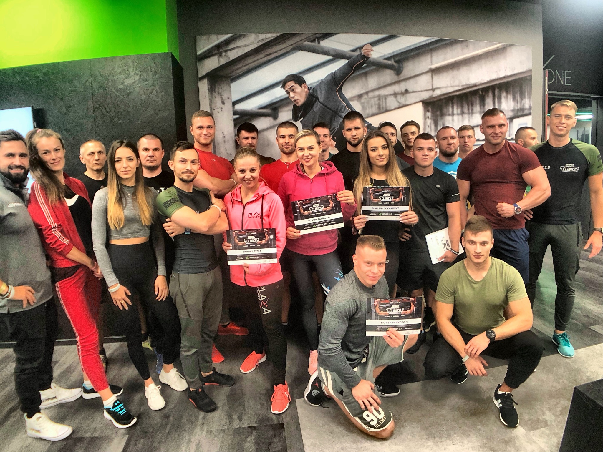 Grupa uśmiechniętych osób w sportowych strojach pozuje z certyfikatami ukończenia kursu trenerskiego w siłowni. W tle grafika z ćwiczącym mężczyzną.