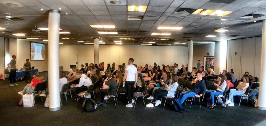 Szeroki kadr sali konferencyjnej wypełnionej uczestnikami szkolenia. Na pierwszym planie siedzący ludzie, w tle prelegenci przy stole i ekran z prezentacją.