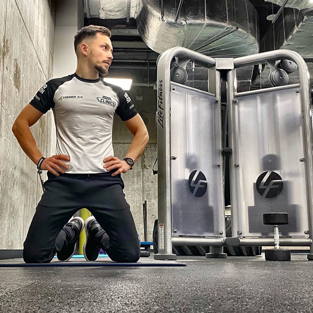 Mężczyzna w sportowej koszulce klęczy obok maszyny Life Fitness w siłowni, spoglądając w bok. Widoczny betonowy mur i instalacja pod sufitem.